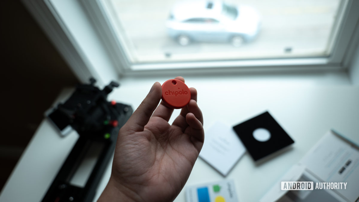 A picture of the Chipolo One tracker held above its packaging in front of a window