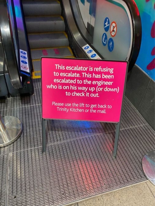 Pink and black sign at the bottom of an escalator which says "This escalator is refusing to escalate. This has been escalated to the engineer who is on his way up (or down) to check it out. 