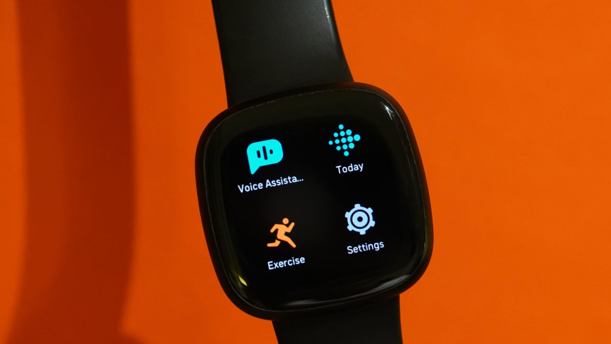 A Fitbit Versa 3 displays its apps in front of an orange background.