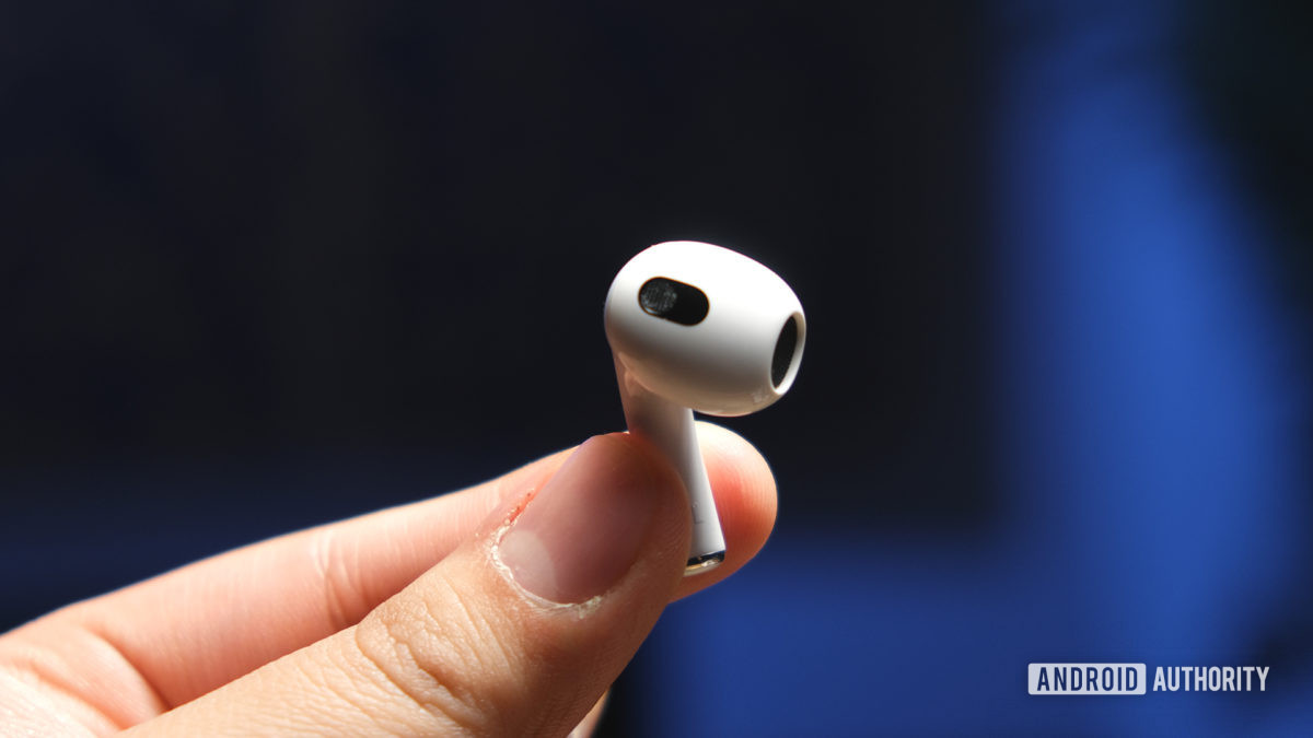 A hand holds a Apple AirPods (3rd generation) earbud by the stem to reveal the open-type fit and embedded sensors.