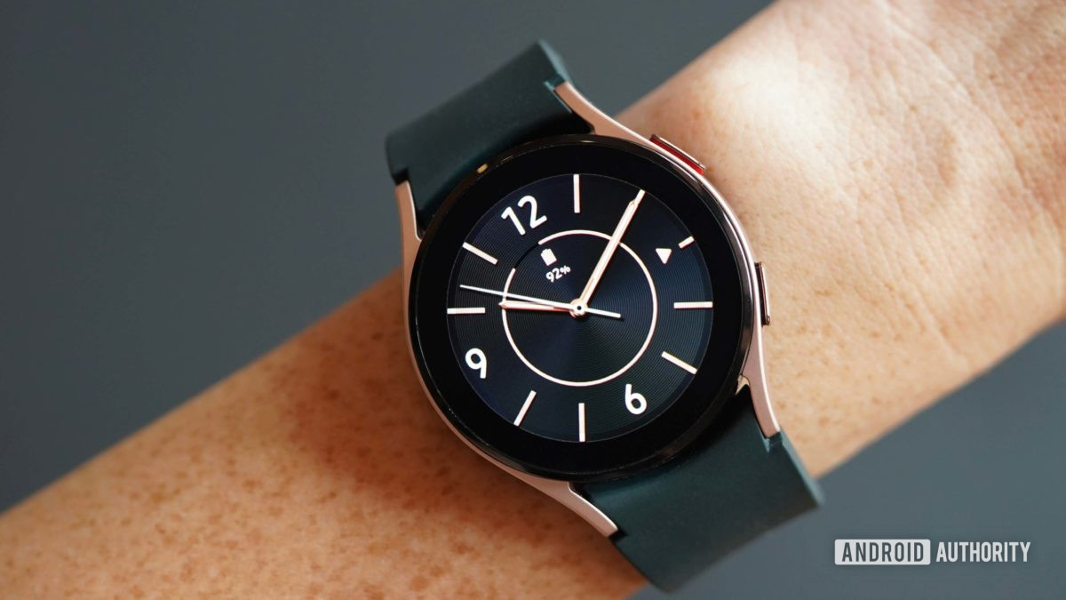 A Galaxy Watch 4 on a female's wrists displays a black and rose gold analog watch face.