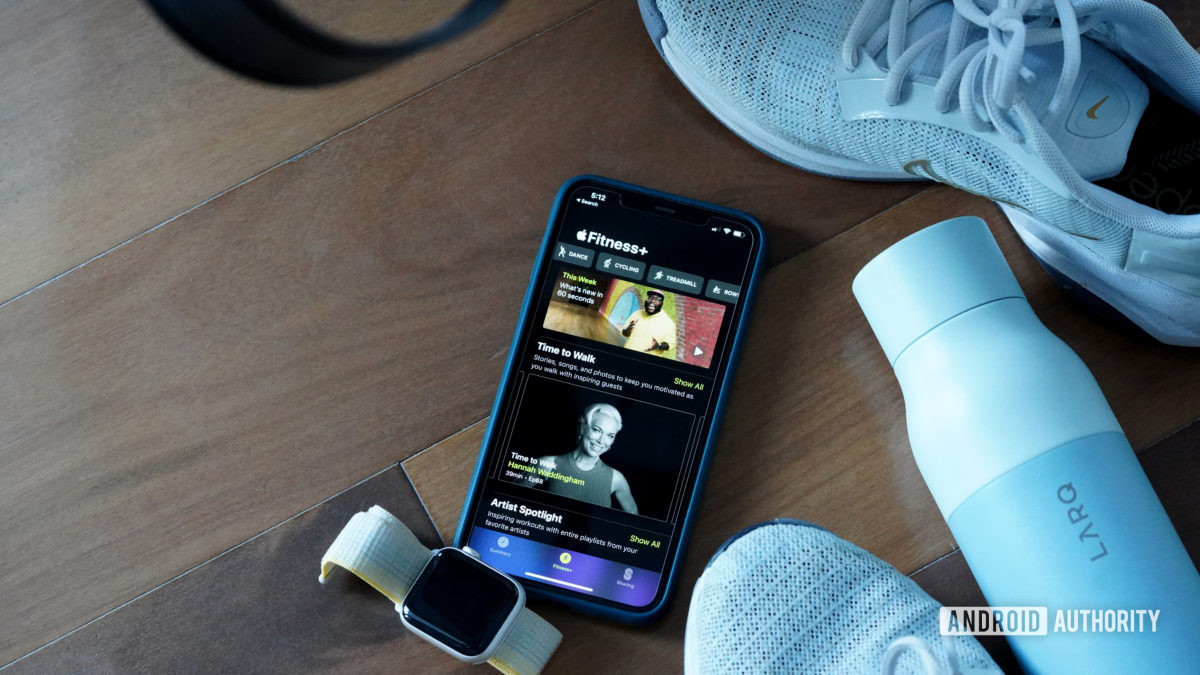 An iPhone displaying Apple Fitness Plus rests on a studio floor among tennis shoes, a water bottle, and an Apple Watch SE 2.