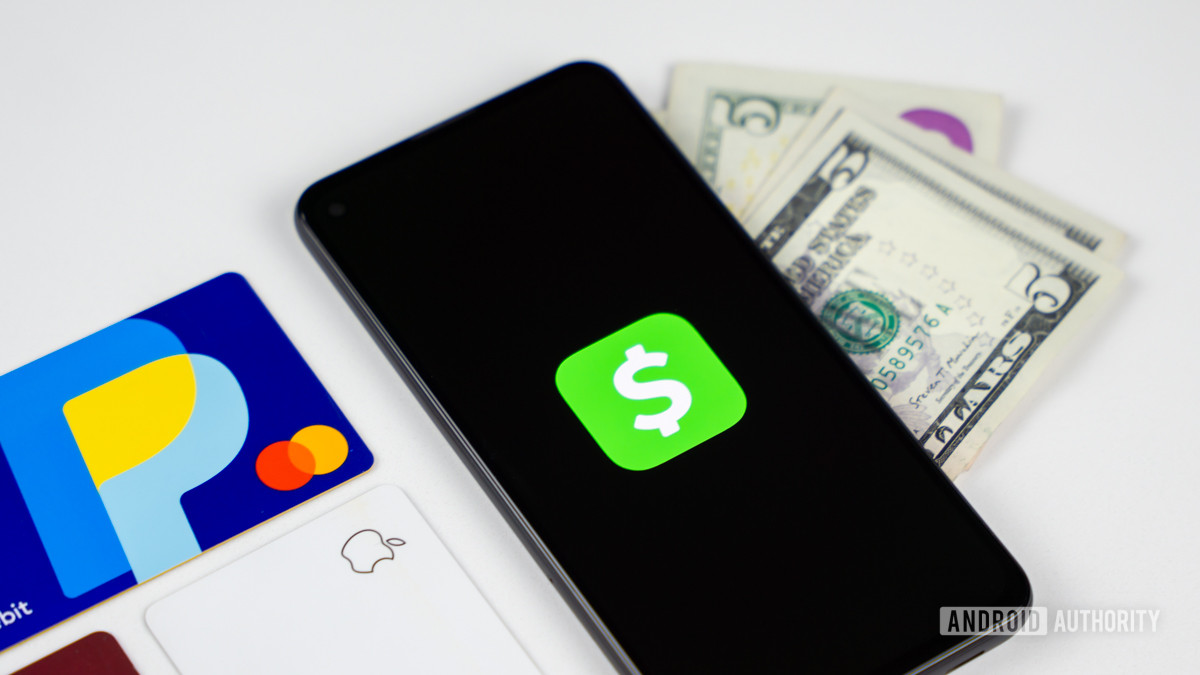 A stock photo of a smartphone showing the Cash App logo on its screen. It lies on top of some cash and next to various gift and debit cards.