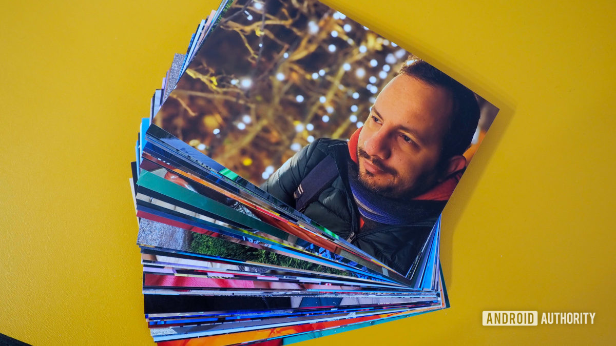 A bunch of photo prints, ordered from Google Photos prints, on a yellow background. The top one shows a smiling man with Christmas lights in the background.