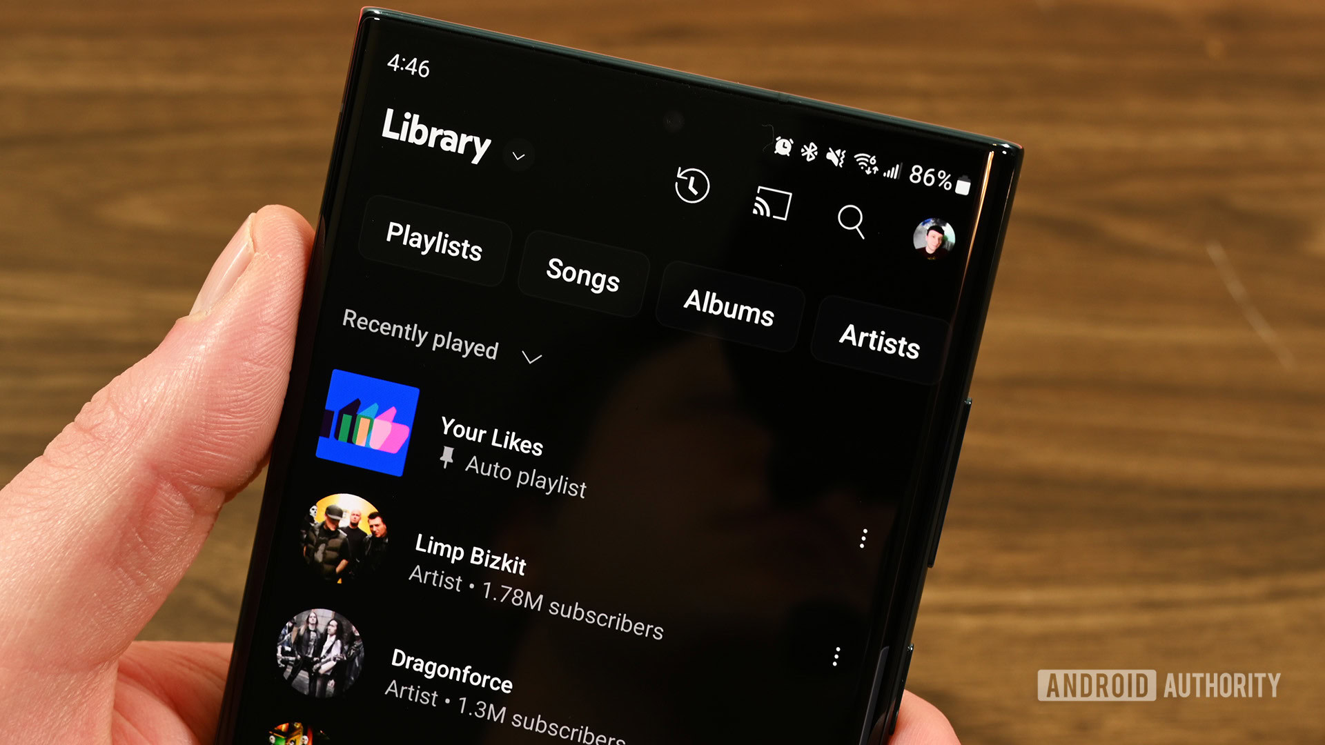 Smartphone in hand on a wooden background with YouTube Music's Library page open on the screen.