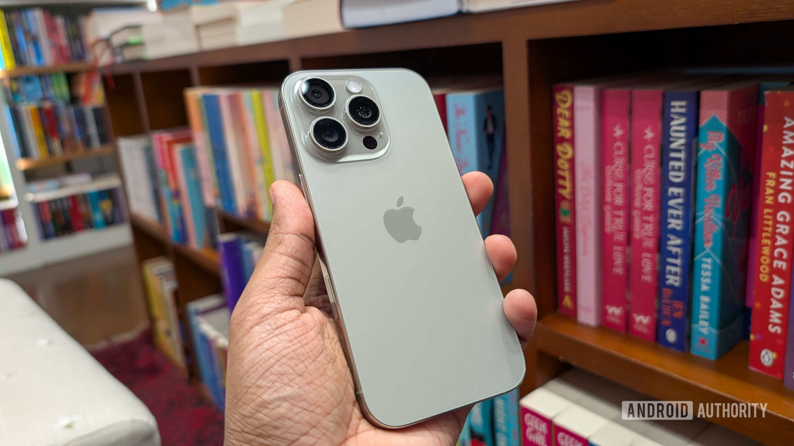 Apple iPhone 16 Pro in Natural Titanium color held out in hand, against the backdrop of books