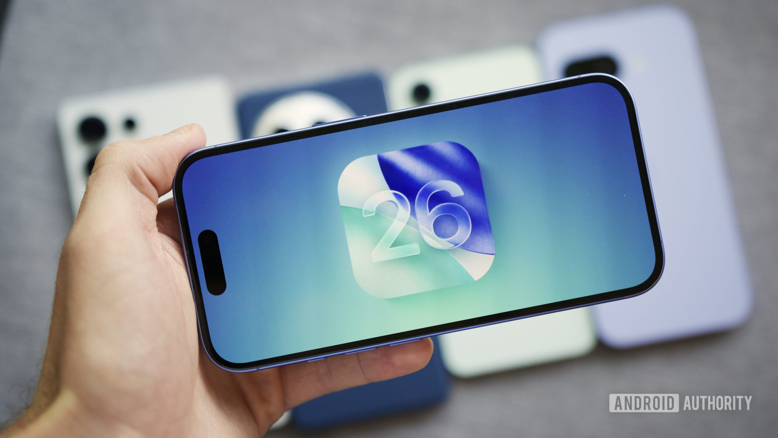 iOS 26 logo on an iPhone, with Android phones in the background.