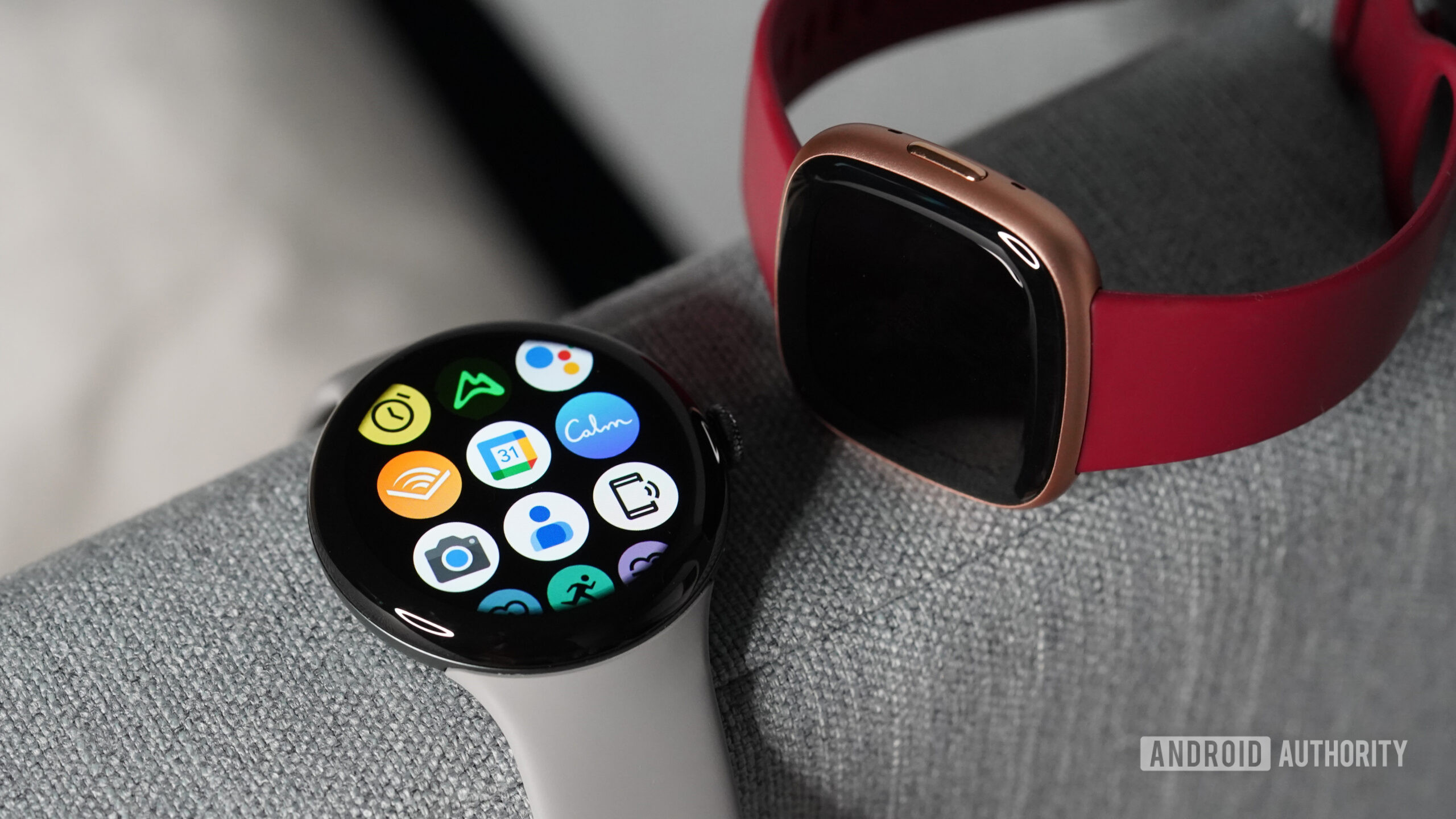 A Pixel Watch displays its app library next to a Fitbit Versa 4.